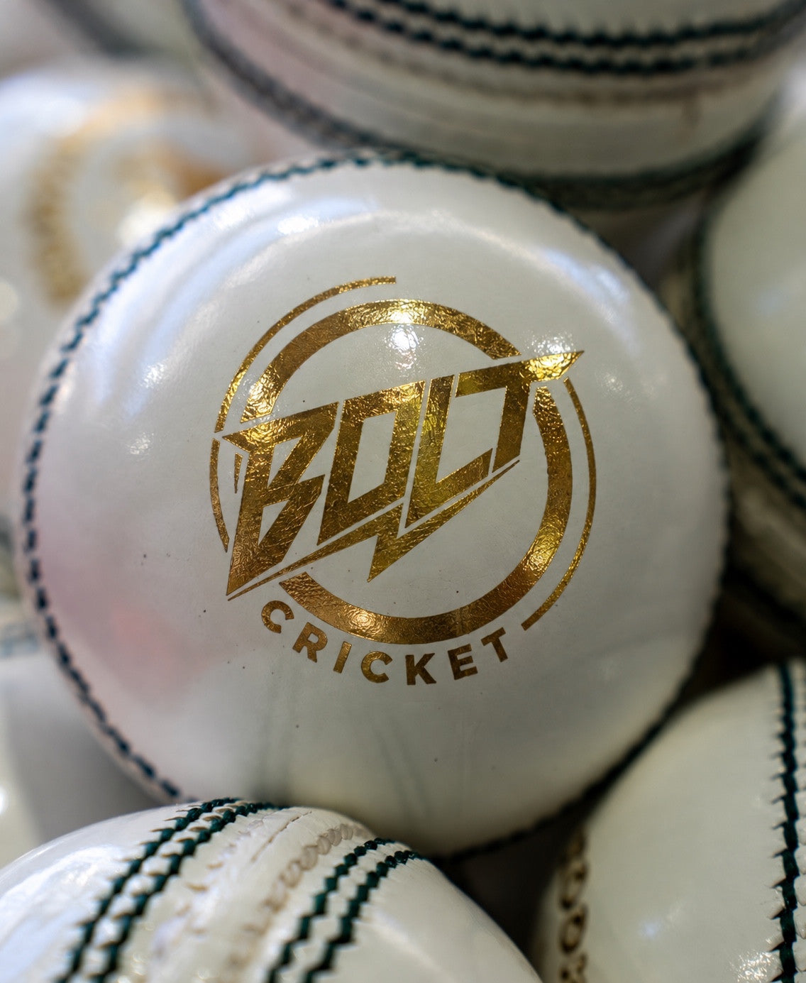 White Bolt Enduro cricket ball with gold Bolt Cricket logo and dark green seam, among other white balls